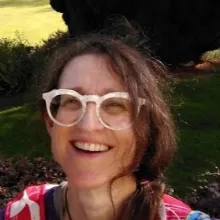 Face profile of Mary Bairstow wearing large pale pink glasses , tied back brown hair and a smile